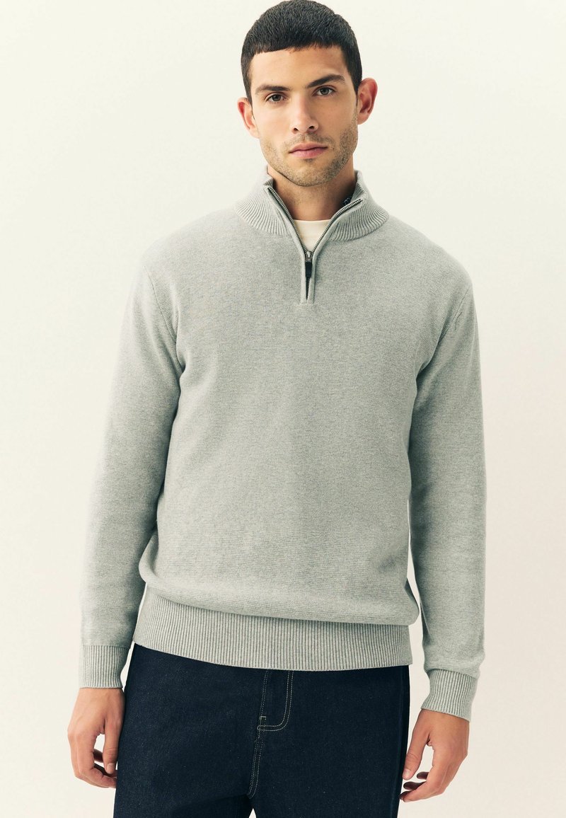 Man in een lichtgrijze quarter-zip trui en donkere jeans tegen een effen lichte achtergrond, kijkend naar voren met een neutrale uitdrukking.
