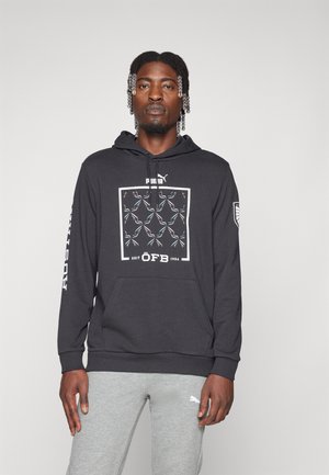 Schwarzer Hoodie mit einem Frontgrafik von Zweigen in Weiß und Teal, ausgestattet mit einer Kängurutasche und gerippten Ärmeln. Getragen mit grauen Hosen.