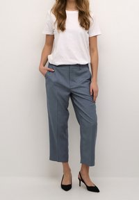 Kvinna iklädd en vit kortärmad t-shirt, blå randiga kortbyxor och svarta spetsiga högklackade skor, stående mot en vit vägg.