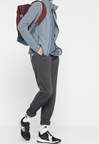 Vaaleansininen fleece-takki, harmaat kapenevat housut ja mustat juoksukengät valkoisin yksityiskohdin. Punainen ja sininen reppu, näkyvät vetoketjut ja hihansuut.