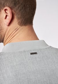 La maglietta polo grigia chiara presenta un colletto a costine e un tessuto testurizzato. Un piccolo marchio scuro è visibile sul retro, vicino al collo.