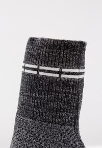 Chaussette tricotée noire et grise avec une texture côtelée, présentant des accents de rayures blanches au niveau de la cheville. Vue en gros plan mettant en avant les détails du design.