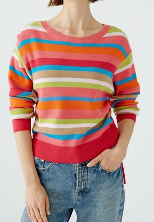 Persona che indossa un maglione a righe multicolori con le maniche arrotolate e jeans in denim blu, mano appoggiata nella tasca, su sfondo semplice.