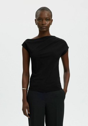 Donna con capelli corti che indossa una canotta asimmetrica nera senza maniche e pantaloni neri, in piedi con una mano in tasca su sfondo bianco.