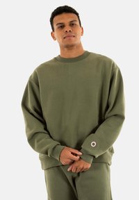 Sudadera verde hecha de tejido suave, con un ajuste holgado, cuello redondo y puños de canalé con un distintivo detalle de botón.