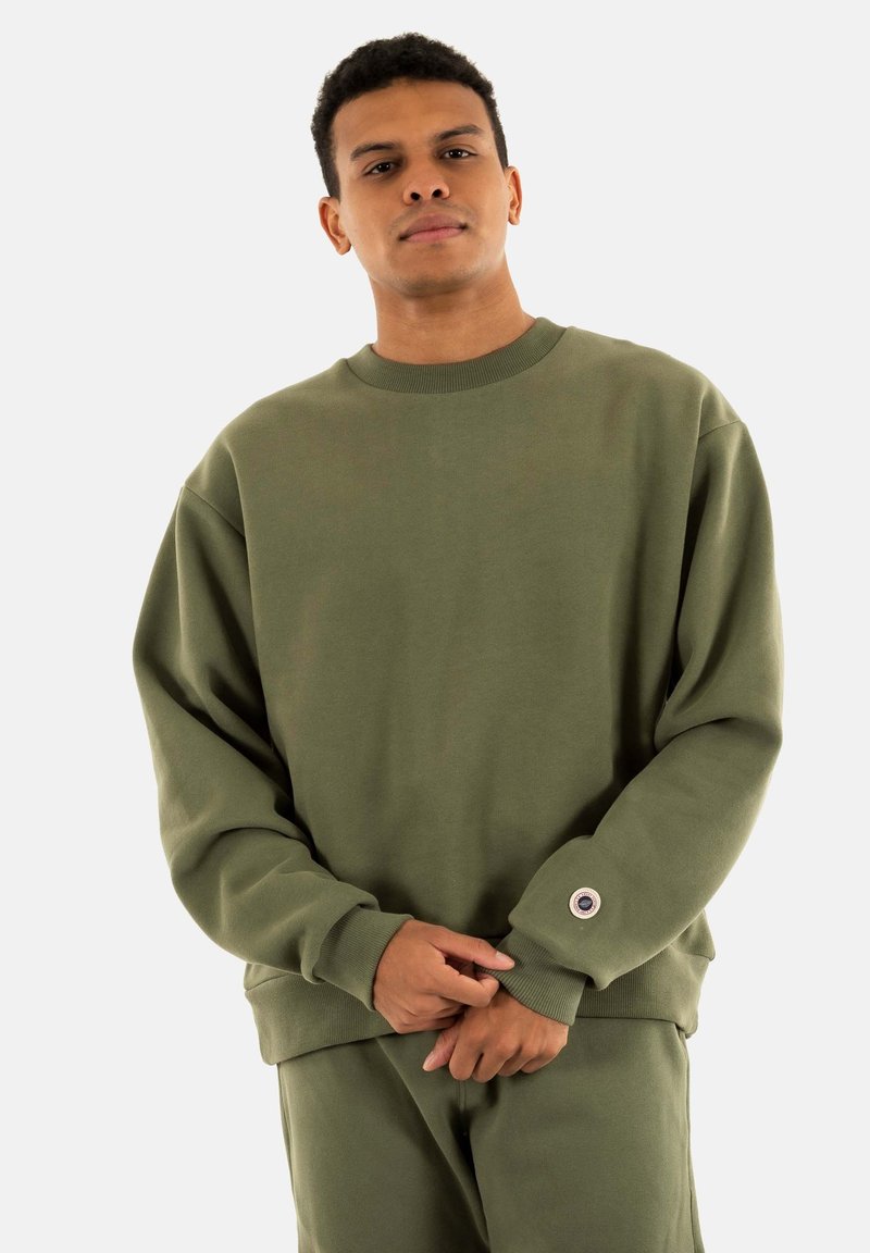 Sudadera verde hecha de tejido suave, con un ajuste holgado, cuello redondo y puños de canalé con un distintivo detalle de botón.