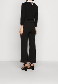 Svart långärmad topp med vågig vit krage och detalj med nyckelhål bak, tillsammans med svarta utsvängda byxor och klumpiga sandaler med klack.