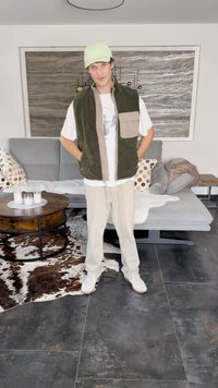 Fleece vest in olive green and beige, paired with a white t-shirt and light beige pants. Cap is light green. Setting includes a patterned rug.