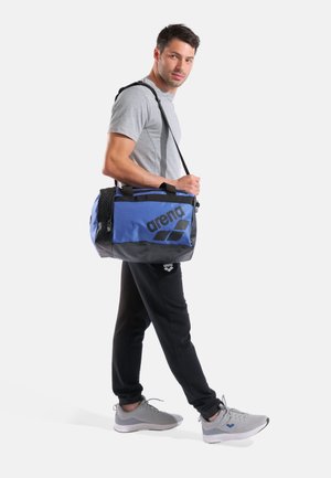 Uomo con maglietta grigia, pantaloni neri e scarpe da ginnastica grigie che porta una borsa sportiva Arena blu e nera sulla spalla.