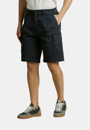 Uomo che indossa pantaloncini cargo neri con tasca laterale e sneaker casual bicolore, in piedi con una mano in tasca su sfondo bianco.