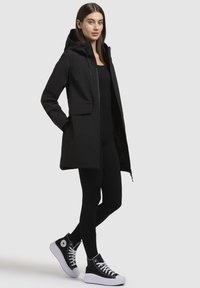 Black, hooded parka coat with a zip front, featuring two front pockets. Paired with black leggings and black high-top sneakers with white soles.