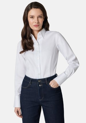 Kobieta o długich brązowych włosach, ubrana w białą koszulę zapinaną na guziki i ciemnoniebieskie jeansy z wysokim stanem, z jedną ręką w kieszeni, neutralne tło.