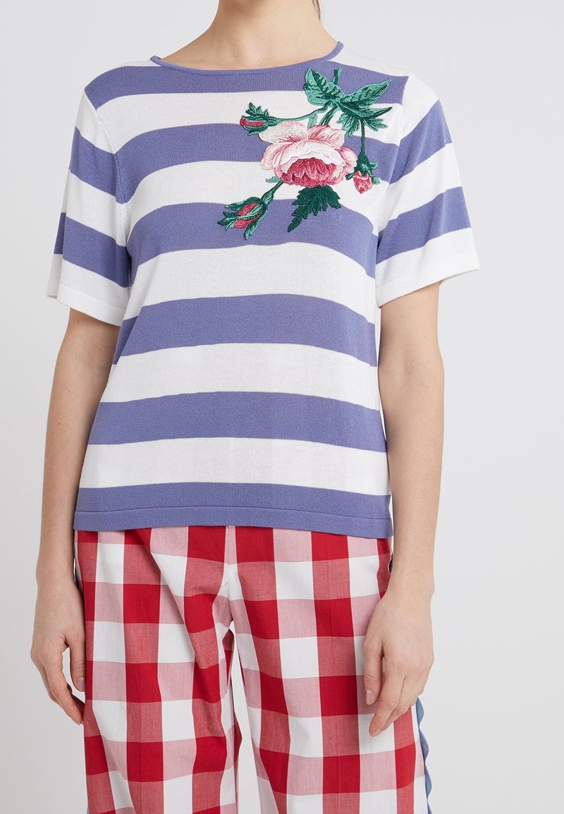 Camiseta de rayas en púrpura y blanco, con un detalle de bordado floral en el pecho. Hecha de tela ligera y con mangas cortas.