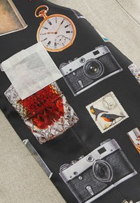 Schwarzer Stoff mit Vintage-Objekten: einer goldenen Taschenuhr, Kameras, einem Kristall-Whiskykaraffe mit roliquidem Inhalt und einer Briefmarke.