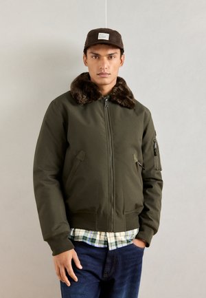 Chaqueta bomber verde oliva con cuello de piel sintética, cierre frontal con cremallera y bolsillos laterales. Llevada sobre una camisa de cuadros y jeans de mezclilla.