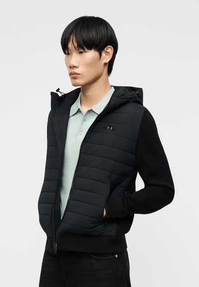 Νέος άνδρας που φοράει μαύρο καπιτονέ μπουφάν με κουκούλα πάνω από ανοιχτό πράσινο πόλο πουκάμισο, στέκεται με τα χέρια στις τσέπες του μπουφάν.