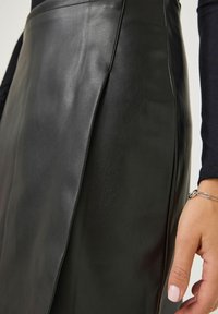 Gros plan sur une jupe en cuir noire avec une fente sur le côté, portée avec un haut à manches longues noir et un bracelet en chaîne argenté au poignet.