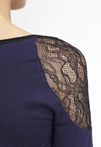 Lila Pullover mit solidem Strickkörper und schwarzen Spitzensäumen. Verfügt über ein zartes Blumenmuster und einen kontrastierenden schwarzen Halsauschnitt.