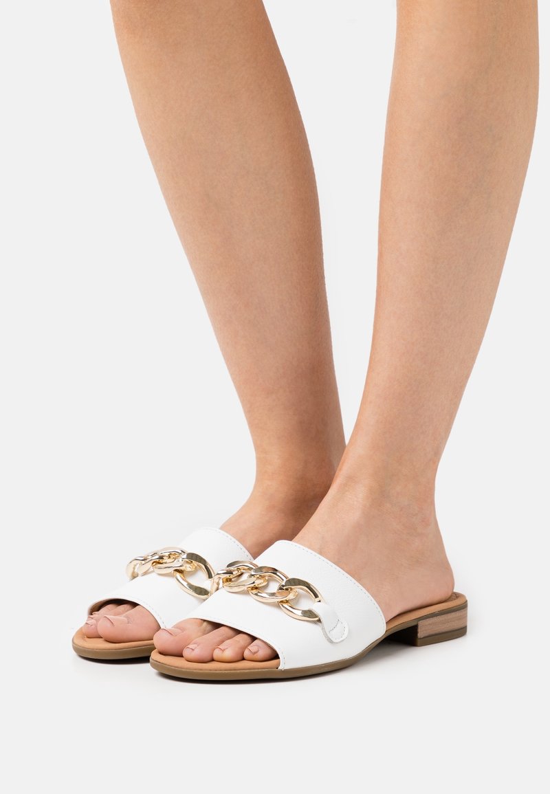 Pieds dans des sandales blanches à enfiler avec un large détail chaîne dorée à l'avant et un petit talon en bois sur fond uni.