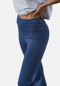 Niebieskie elastyczne spodnie z wysokim stanem, gładką fakturą i dopasowanym krojem, prezentowane przez modelkę w białej bluzce na neutralnym tle.