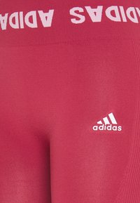 Leggings rouges avec une finition texturée, présentant une taille large marquée par "ADIDAS" en blanc et un petit logo blanc sur la jambe inférieure.
