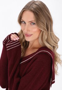 Maglione bordeaux con accenti a strisce bianche, texture a coste, colletto ampio e maniche lunghe. Il materiale sembra morbido e lavorato a maglia.