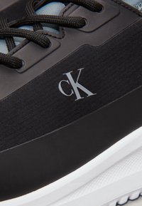 Zapatilla negra con tejido texturizado, suela de goma blanca, cordones negros y pequeño logo "CK" plata en el lateral. Presenta acentos de plástico liso.