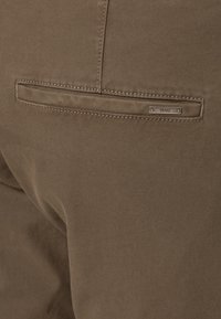 Gros plan sur un tissu marron d'une poche arrière d'un pantalon avec une étiquette de marque métallique Hugo Boss cousue au-dessus de l'ouverture de la poche.