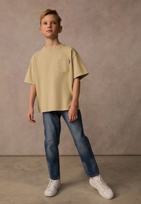 Beige t-shirt med kort ärm och ficka, i kombination med blå raka jeans och vita träningsskor. En enkel, avslappnad look med minimala detaljer.