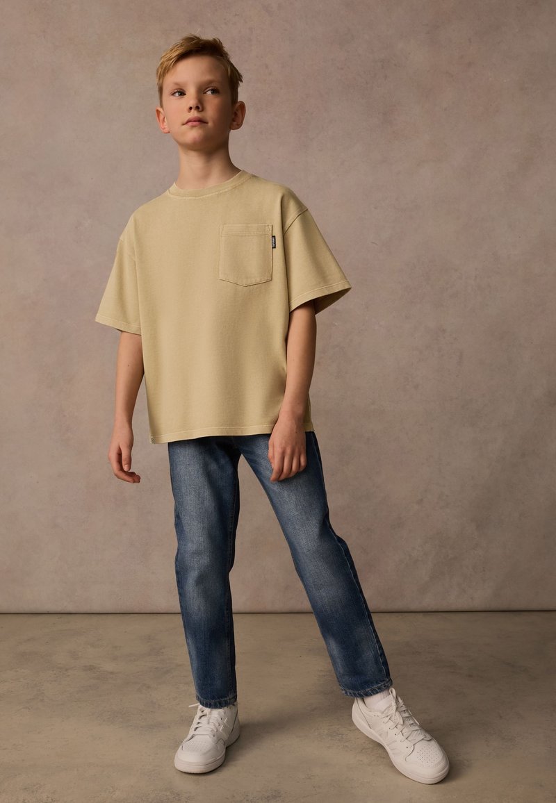 Beige t-shirt med kort ärm och ficka, i kombination med blå raka jeans och vita träningsskor. En enkel, avslappnad look med minimala detaljer.