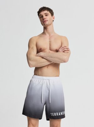 Giovane uomo in piedi con sicurezza, braccia incrociate, indossa pantaloncini da bagno con sfumatura dal nero al bianco e logo "TERRANOVA".