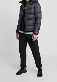 Giacca a vento nera con cuciture orizzontali e cappuccio, indossata sopra un felpa nera e una maglietta grigia, abbinata a pantaloni cargo neri e sneakers.