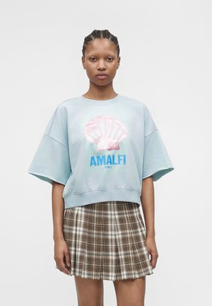 Ung kvinde med flettet hår iført en lyseblå oversized skjorte med lyserødt skaldyr og teksten "AMALFI ITALY", kombineret med en brun ternet nederdel.