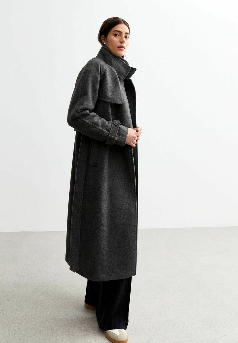 Cappotto lungo in lana grigia con colletto alto, tasche laterali e polsini abbottonati. Texture liscia con silhouette dritta, indossato sopra pantaloni scuri.