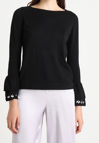 Zwarte gebreide trui met een bootcut halslijn, uitlopende mouwen en witte bloemenborduursels op de boorden. Gecombineerd met lichtgekleurde broek.