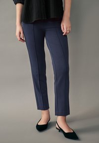 Pantaloni navy, croiți pe corp, cu cusături verticale, textură netedă și lungime scurtă, asortați cu pantofi cu toc de culoare verde închis.
