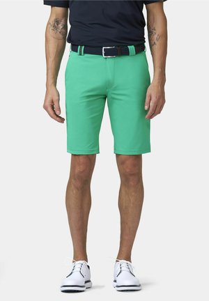 Mann trägt leuchtend grüne knielange Shorts, weiße Schnürschuhe, einen dunklen Gürtel und ein dunkles kurzärmeliges Hemd, steht mit entspannten Armen.