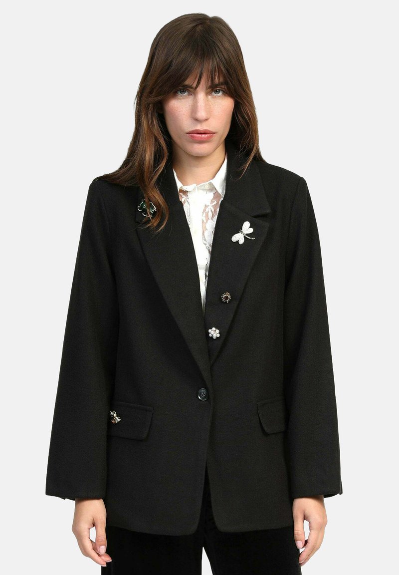 Femme portant un blazer noir avec des épingles décoratives sur un blouse en dentelle blanche, se tenant devant un fond clair.