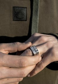 Silberner Ring mit erhabenen rechteckigen Mustern, mit einer glatten, polierten Oberfläche. Die Hände ruhen auf einem strukturierten, olivgrünen Stoff.