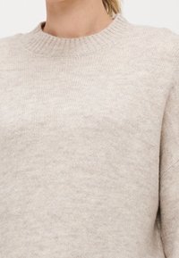 Beige pletený svetr s vroubkovaným kulatým výstřihem a volným střihem. Texturovaná látka vykazuje jemné variace v barvě a měkkosti.