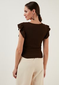 Top marrone a coste con maniche corte e volants, abbinato a pantaloni color crema. Il tessuto ha un aspetto texturizzato e una silhouette aderente.