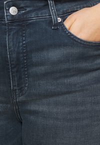 Jeans de mezclilla oscura con una textura suave, que presentan un bolsillo visible y un botón plateado con el logo 'CK' en la cintura.