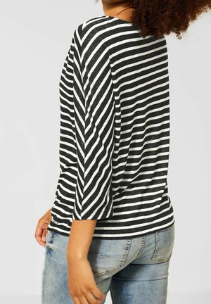 Persona vistiendo una camiseta de manga larga a rayas blancas y negras y pantalones vaqueros azul claro, mostrada de espaldas con cabello rizado.