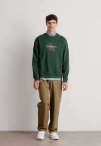 Sweatshirt verde com o logótipo "Calvin Klein", combinado com calças beges e sapatos brancos; inclui uma camisa de colarinho azul clara por baixo.