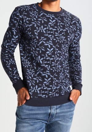 Homme portant un pull bleu marine avec des motifs de feuilles bleu clair et un jean bleu, les mains dans les poches.