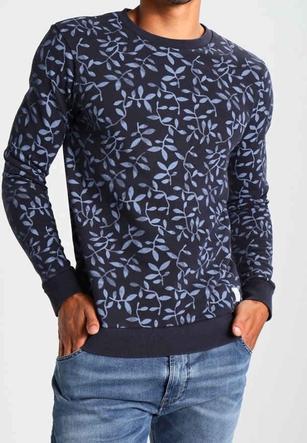 Homme portant un pull bleu marine avec des motifs de feuilles bleu clair et un jean bleu, les mains dans les poches.