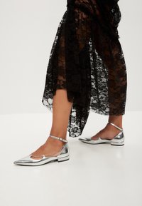 Silberne metallische flache Schuhe mit spitzen Zehen und Knöchelriemen, vor einem transparenten schwarzen Spitzenrock mit floralen Mustern.