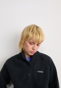 Giovane con capelli biondi corti che indossa un pullover in pile nero Columbia, che guarda verso il basso su uno sfondo bianco semplice.