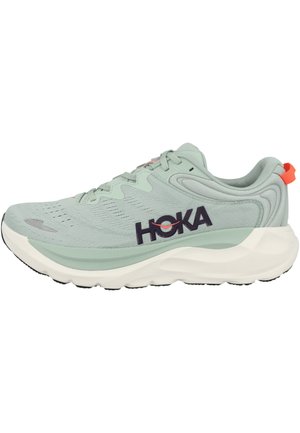 Lichtgroene hardloopschoen met dikke witte zool, zwart "HOKA" logo aan de zijkant, oranje hieltab en ademend mesh bovenwerk.