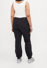 Svarta cargobyxor med elastiska muddar, fickor och smal midja. Kombinerat med en vit ärmlös topp och grå sportskor.